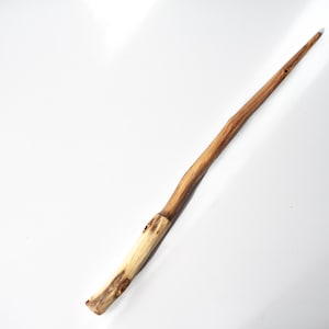 Pode incluir: Uma varinha de madeira longa e fina com um acabamento natural castanho claro e veios de madeira visíveis. A secção do cabo é mais espessa e clara, transformando-se num eixo cónico castanho escuro com uma textura lisa e polida.
