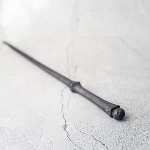 May include: A black wooden wand with a rounded tip. The wand is lying on a light grey surface.