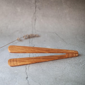 Puede incluir: Pinzas de cocina de madera con un acabado de madera natural. Las pinzas están hechas de una sola pieza de madera y tienen un diseño simple.
