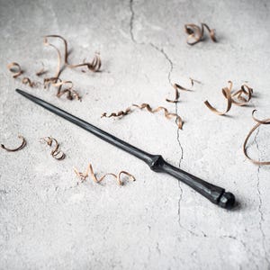 May include: A black wooden wand with a rounded tip, lying on a gray surface with brown wood shavings scattered around it.