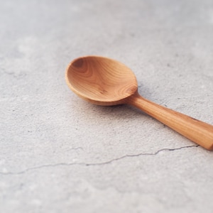 Könnte beinhalten: Ein Holzlöffel mit einer abgerundeten Schüssel und einem langen, schlanken Griff. Der Löffel ist aus hellem Holz gefertigt, das die natürliche Maserung zeigt. Der Löffel liegt vor einem grauen, strukturierten Hintergrund.