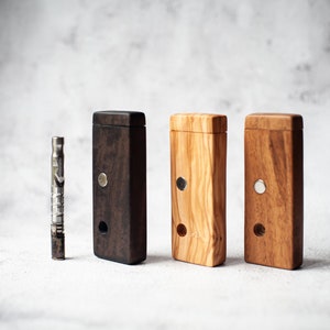 Puede incluir: Tres cajas de madera con cierres magnéticos, una negra, una marrón claro y una marrón oscuro. Una pipa de metal plateado está delante de las cajas.