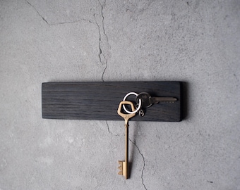 May include: A black wooden key holder with a set of keys attached to it. The key holder is mounted on a gray wall.