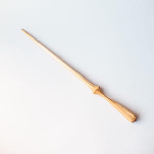Puede incluir: Una varita de madera de color claro con un eje liso y cónico que termina en una punta. La sección del mango es ligeramente más gruesa, con un collar distintivo en forma de diamante que separa la empuñadura del cuerpo largo y delgado.