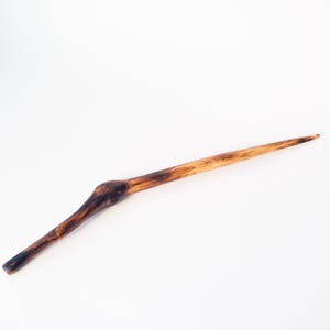 Puede incluir: Un palillo para el pelo largo y delgado hecho de madera pulida, con un patrón de veta natural con vetas de color marrón oscuro y bronceado claro. El diseño incluye una sección ligeramente más gruesa y redondeada cerca de la base.