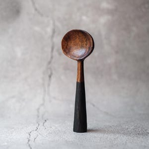 Peut inclure: Une cuillère en bois faite à la main, avec un bol rond et un long manche. Le bol est brun chaud, le manche est noir. La cuillère est posée sur un fond gris neutre.