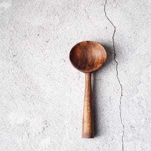 Peut inclure: Une cuillère en bois avec un bol arrondi et un long manche fin. La cuillère est faite d'un bois foncé et riche et est posée sur un fond texturé gris clair. La cuillère est un ustensile de cuisine.