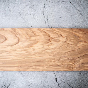 Könnte beinhalten: Ein rechteckiges Schneidebrett aus Holz mit natürlicher Holzmaserung. Das Brett ist aus Olivenholz und hat eine hellbraune Farbe.