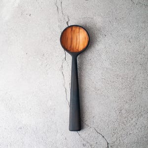 Könnte beinhalten: Ein handgefertigter Holzlöffel mit einem dunklen, mattschwarzen Griff und einer Schüssel in Naturholzoptik. Der Löffel hat einen langen, schlanken Griff und eine abgerundete Schüssel, ideal zum Servieren oder Kochen.