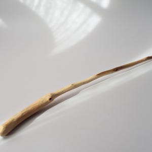 Könnte beinhalten: Ein handgeschnitzter Zauberstab aus Holz mit einer hellbraunen, natürlichen Oberfläche. Das Objekt verfügt über einen dickeren, zylindrischen Griff, der sich allmählich zu einer schlanken, spitzen Endung verjüngt und die organische Textur sowie die Maserung des Holzes zeigt.