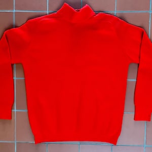 Puede incluir: Un jersey rojo brillante de cuello alto. El jersey de manga larga está hecho de un material de punto. El cuello y los puños son de un tono rojo más oscuro. El jersey se muestra sobre una superficie de baldosas marrones.