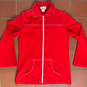 May include: A vibrant red jacket featuring a white zip and white stitching. The jacket has a collar, two front pockets, and long, flared sleeves. The garment is a solid colour and appears to be made of a smooth fabric.