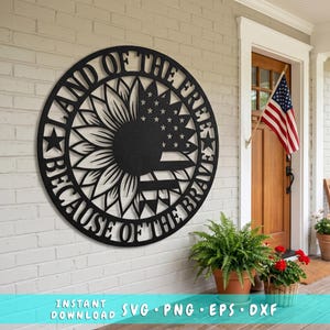 Può includere: Opera d'arte da parete in metallo nero con un design a girasole e la bandiera americana. Il testo recita "Land of the Free Because of the Brave". L'opera è esposta su un muro di mattoni bianchi. È visibile anche una piccola bandiera americana.