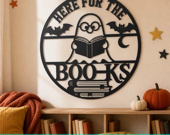 May include: A circular black wall sign featuring a ghost wearing glasses and reading an open book. The text reads "HERE FOR THE BOO-KS". Two bats and a crescent moon are integrated into the design, with a stack of books at the bottom.