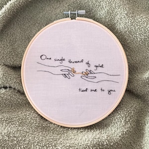May include: Embroidered hoop art with a pink fabric background and a black thread design of two hands connected by a gold thread. The text reads "One single thread of gold tied me to you."