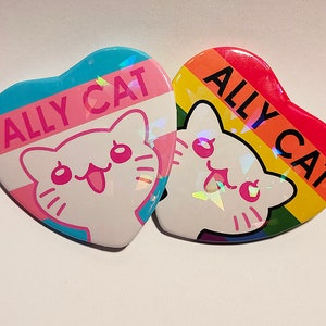 Könnte beinhalten: Zwei herzförmige Anstecknadeln mit einer weißen Katzenillustration und dem Text "ALLY CAT" auf jeder. Eine Nadel hat ein Transgender-Pride-Flaggen-Design und die andere ein Regenbogen-Pride-Flaggen-Design.