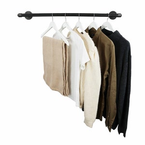 Könnte beinhalten: Ein schwarzer Metall-Kleiderständer mit fünf weißen Kleiderbügeln, an denen verschiedene Kleidungsstücke hängen, darunter ein beiger Pullover, ein weißes T-Shirt, ein cremefarbener Pullover, ein braunes Hemd und ein dunkelgrauer Pullover.