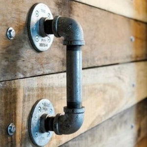 Puede incluir: Una manija de puerta de tubo industrial plateado con dos bridas redondas fijadas a una pared de madera.