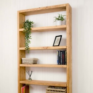 May include: A wooden bookshelf with five shelves. The shelves are empty except for a small potted plant, a framed picture, a wicker basket, a small bottle of essential oils, and a stack of books. The bookshelf is made of natural wood and has a rustic look.