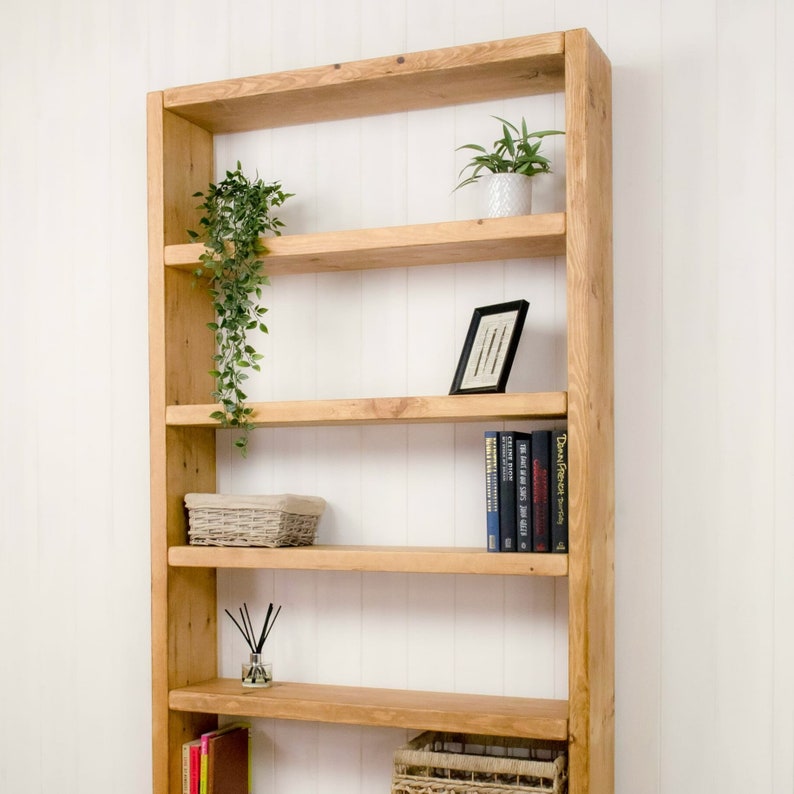 May include: A wooden bookshelf with five shelves. The shelves are empty except for a small potted plant, a framed picture, a wicker basket, a small bottle of essential oils, and a stack of books. The bookshelf is made of natural wood and has a rustic look.