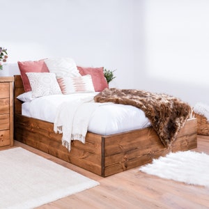 May include: A wooden bed frame with a white mattress and a brown faux fur throw. The bed has a storage compartment underneath the mattress. The bed is decorated with pillows in shades of pink and white.