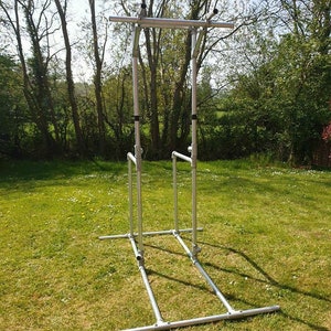 Puede incluir: Una barra de dominadas y estación de flexiones de metal plateado con una barra horizontal y dos juegos de barras verticales. La estación está instalada en un césped.