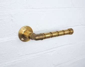 Könnte beinhalten: Ein goldfarbener Toilettenpapierhalter aus Metall mit runder Basis und einem rohrartigen Design. Der Halter ist an einer weißen Backsteinwand montiert.