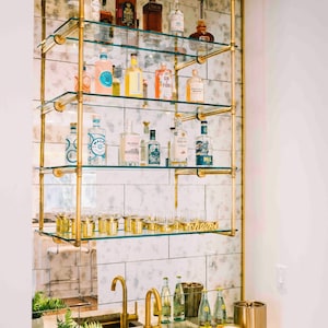 May include: A gold metal bar cart with glass shelves filled with bottles of liquor and glassware. The cart is mounted to a wall with a mirrored tile backsplash. The cart has a gold metal sign that says "cheers".