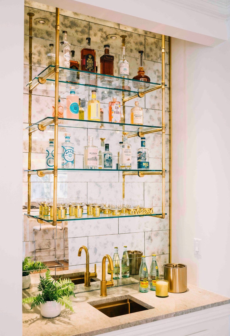 May include: A gold metal bar cart with glass shelves filled with bottles of liquor and glassware. The cart is mounted to a wall with a mirrored tile backsplash. The cart has a gold metal sign that says "cheers".