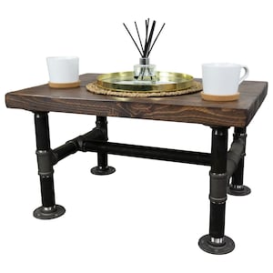 Può includere: Un tavolino da caffè in legno marrone scuro con gambe in metallo nero. Il tavolo ha un vassoio in metallo dorato con un tappetino intrecciato sopra. Due tazze in ceramica bianca sono sul tavolo.