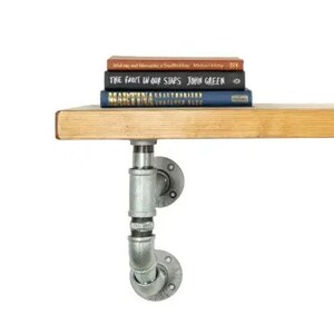 May include: A wooden shelf with a stack of books, supported by industrial-style galvanized steel pipe brackets. The books have various colors and titles. The shelf is made of light-colored wood.