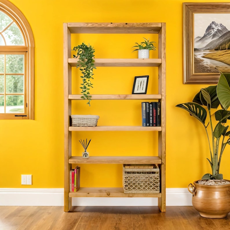 May include: A wooden bookshelf with five shelves, featuring a potted plant, books, a decorative basket, and a small potted plant. The bookshelf is against a yellow wall.