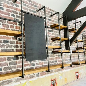 May include: A wall with a brick pattern, featuring a black chalkboard and multiple wooden shelves supported by black metal pipes. The shelves are empty.