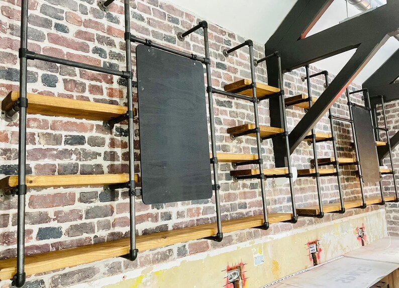 May include: A wall with a brick pattern, featuring a black chalkboard and multiple wooden shelves supported by black metal pipes. The shelves are empty.