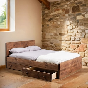 Puede incluir: Un marco de cama de madera con dos cajones, uno de los cuales est&aacute; abierto. La cama est&aacute; hecha con un edred&oacute;n blanco y dos almohadas grises. La cama est&aacute; en una habitaci&oacute;n con una pared de piedra.