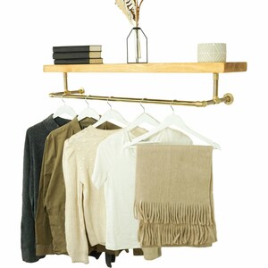 May include: A gold pipe shelf with a wooden top shelf and a gold pipe rod for hanging clothes. There are five white hangers with clothes hanging on them, including a white t-shirt, a beige sweater, a green shirt, a gray shirt, and a beige scarf.