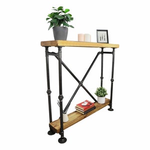 Puede incluir: Consola de estilo industrial con tapa de madera clara y estante inferior. El marco está hecho de tuberías de metal negro, con un soporte en forma de X en los laterales. Decorada con una planta en maceta, velas, un marco de fotos y libros.