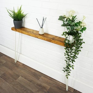 May include: A wooden console table with hairpin legs. The table is stained a dark brown color and has a white brick wall behind it. There are two plants on the table, one with green leaves and one with white flowers.