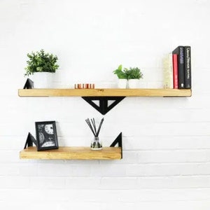 May include: Two wooden shelves with black metal triangular brackets. The top shelf has a small potted plant and a stack of books. The bottom shelf has a framed picture and a small bottle of diffuser sticks.
