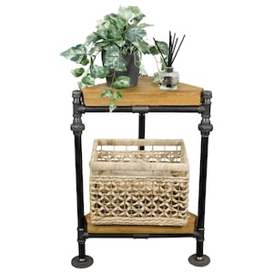 Puede incluir: Una estantería de esquina de estilo industrial de metal negro con dos estantes de madera. El estante superior tiene una planta en maceta con hojas verdes y una botella de vidrio con palitos negros. El estante inferior tiene una cesta tejida.