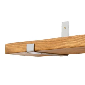 May include: A wooden shelf with a light brown finish, supported by two white metal brackets. The shelf is rectangular and appears to be made of solid wood. The brackets are mounted on the underside and back of the shelf, providing support.