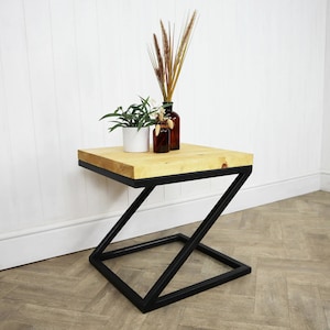 Könnte beinhalten: Ein kleiner Holztisch mit einem schwarzen, Z-förmigen Metallgestell. Der Tisch hat eine helle Holzplatte und ist mit einer Topfpflanze und zwei Glasflaschen dekoriert.