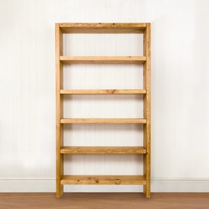 May include: A wooden bookshelf with five shelves. The bookshelf is made of natural wood and has a rustic look. It is standing on a wooden floor in front of a white wall.