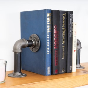 Pode incluir: Suportes para livros de estilo industrial feitos de tubos e acessórios de metal preto. Um está preso a um livro azul, o outro suporta uma pilha de livros. Os suportes para livros adicionam um toque rústico e único a qualquer estante, em cm.