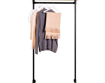 May include: A black metal clothing rack with a wooden top shelf. Three white hangers hold a grey sweater, a white shirt, and a peach-coloured scarf.