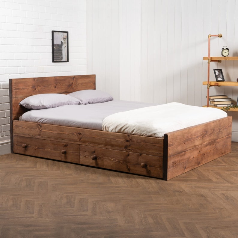 Puede incluir: Un marco de cama de madera con dos cajones y una cabecera. El marco de la cama est&aacute; hecho de madera natural con un tinte marr&oacute;n. La cama est&aacute; hecha con ropa de cama blanca y dos almohadas.