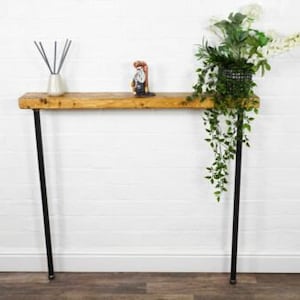 May include: A rustic wooden console table with black metal legs. The table is decorated with a small white vase with sticks, a small figurine, and a hanging plant in a black planter.
