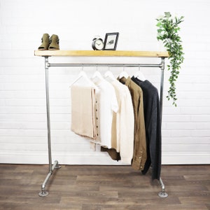 Op de afbeelding: Een witte metalen kledingrek met een houten plank en een hangende stang. Het rek heeft een paar bruine laarzen op de plank, een witte wekker en een ingelijst foto. Er hangen vijf kledingstukken aan het rek, waaronder een wit shirt, een bruine trui, een bruine sjaal en een grijze trui.