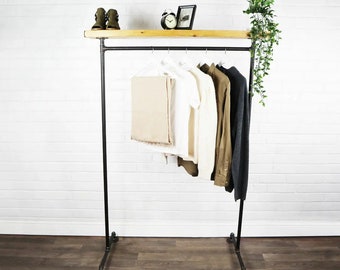 May include: A black metal clothes rail with a wooden shelf. The rail displays a selection of garments: tan trousers, a white t-shirt, a brown shirt, and a dark grey sweater. The shelf holds a small clock, a framed picture, and a pair of boots.