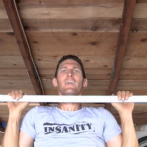 May include: A person doing a pull-up on a white PVC pipe pull-up bar mounted to the ceiling. The bar is attached to black metal pipes. The person is wearing a grey t-shirt with the word "INSANITY" printed on it.